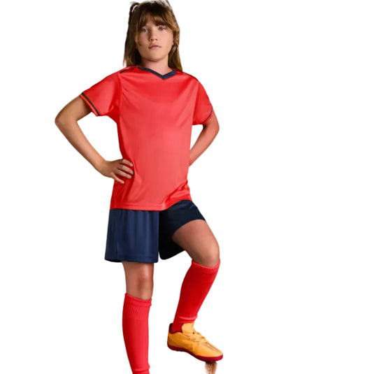 A young girl modeling a red short-sleeve soccer jersey with navy trim, navy shorts, red socks, and yellow soccer cleats, standing on a soccer ball against a solid blue studio background.