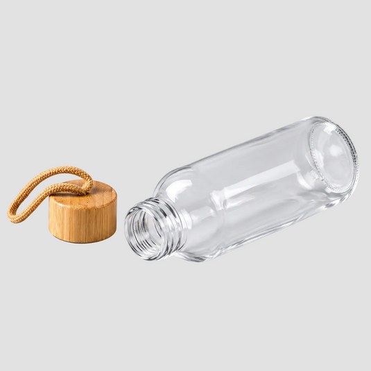 A clear glass bottle with an open neck, shown lying on its side on a light gray background, next to a round bamboo cap with a tan woven loop.