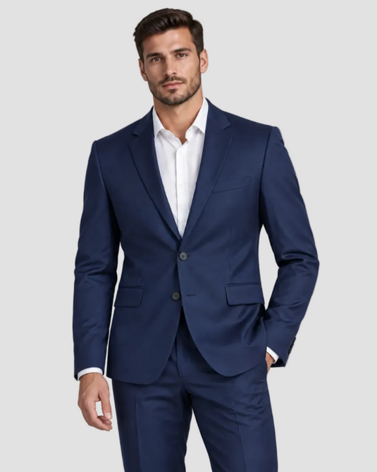 A man wearing a navy blue two-piece suit with a white dress shirt, standing against a solid light gray studio background.
