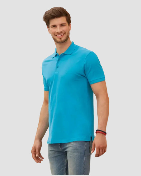 A man wearing a bright turquoise blue short-sleeve polo shirt and blue jeans, standing against a beige stone wall.