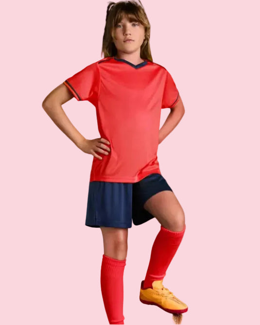 A young girl modeling a red short-sleeve soccer jersey with navy trim, navy shorts, red socks, and yellow soccer cleats, standing on a soccer ball against a solid blue studio background.