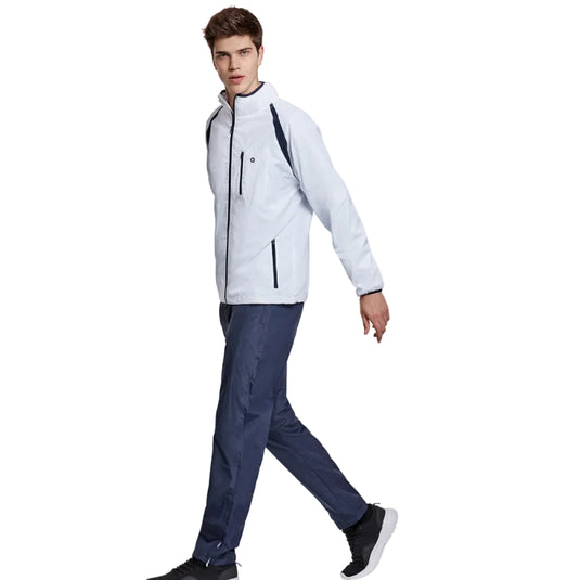 A man wearing a light gray and navy blue athletic jacket, dark blue pants, and black sneakers, walking against a plain light gray studio background.