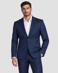 A man wearing a navy blue two-piece suit with a white dress shirt, standing against a solid light gray studio background.