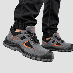 A pair of gray safety work boots with black and orange accents, shown on a person wearing black pants against a gray studio background.