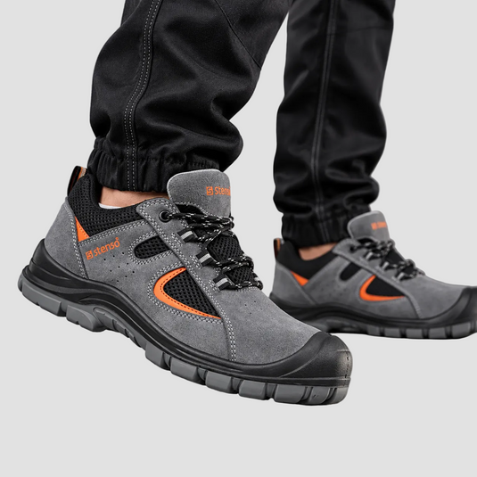 A pair of gray safety work boots with black and orange accents, shown on a person wearing black pants against a gray studio background.