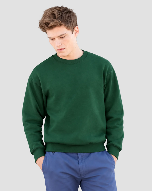 A man wearing a dark green crewneck sweatshirt and blue pants, standing against a solid gray studio background.