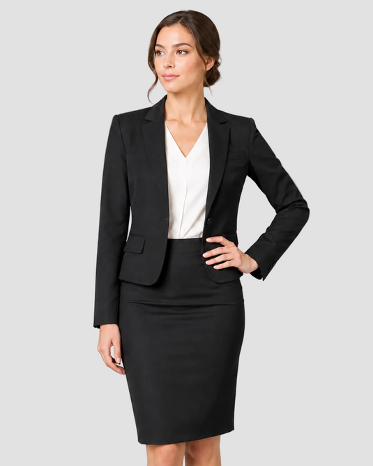 A woman modeling a black two-piece skirt suit consisting of a tailored blazer and a matching pencil skirt, paired with a white V-neck blouse, against a solid gray studio background.