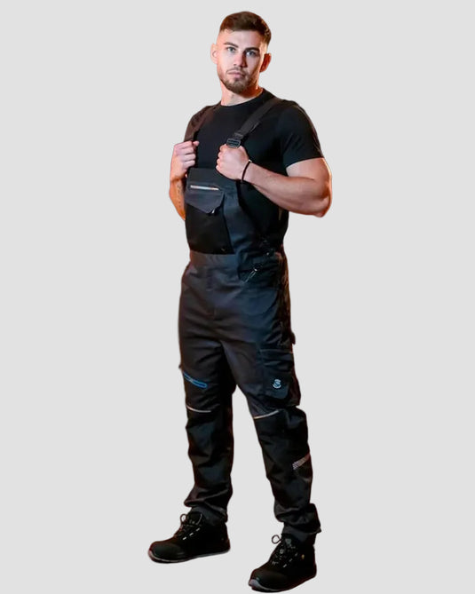 A man wearing black overalls over a black t-shirt and black boots, standing in a brick archway corridor with warm lighting.